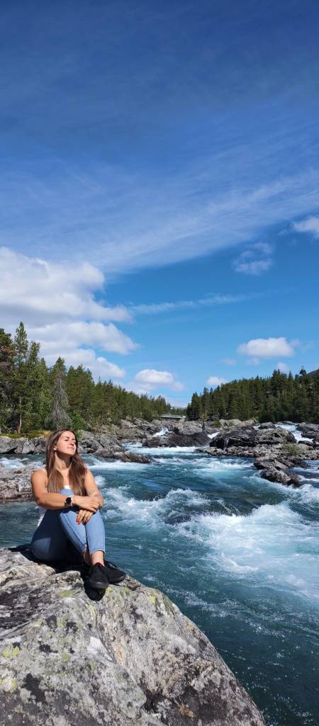 Osoba siedząca na głazie nad rzeka, otoczona zielonymi drzewami, z niebieskim niebem w tle.